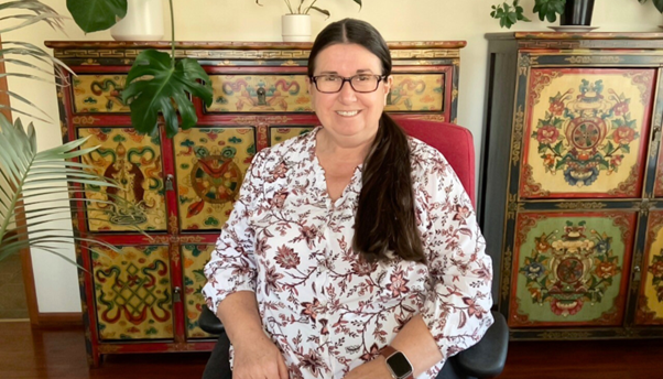 Sharon is smiling facing the camera, sitting in her red and black office chair. She is wearing a floral-patterned top.