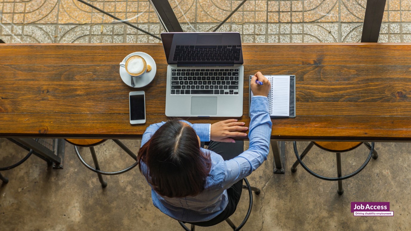 Image description: person writing notes with laptop, coffee and phone, looking from overhead.