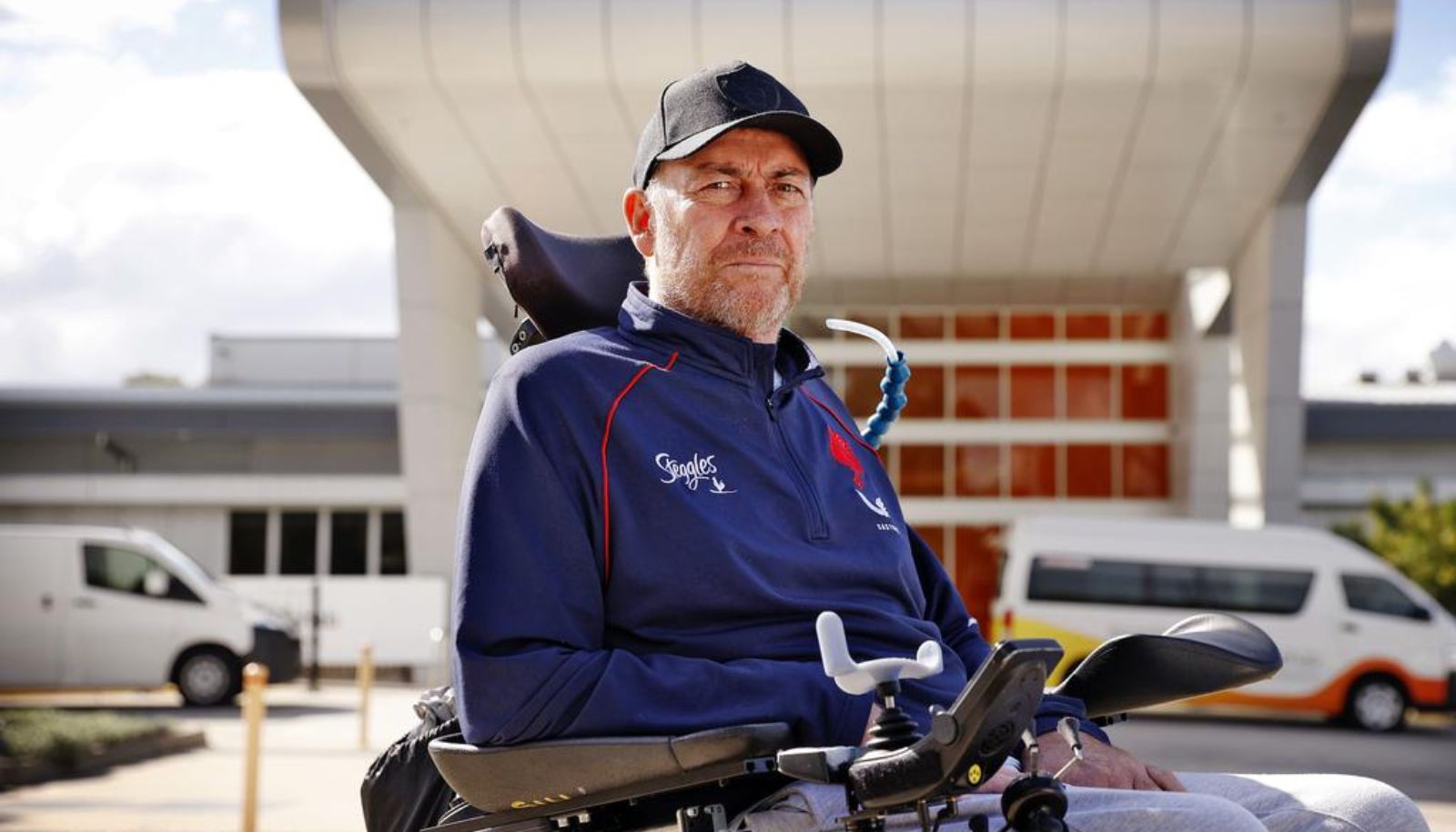 Daniel is wearing a blue jacket and black cap. He is looking at the camera. There is a building and two cars behind him. Photo credit: Daily Telegraph.