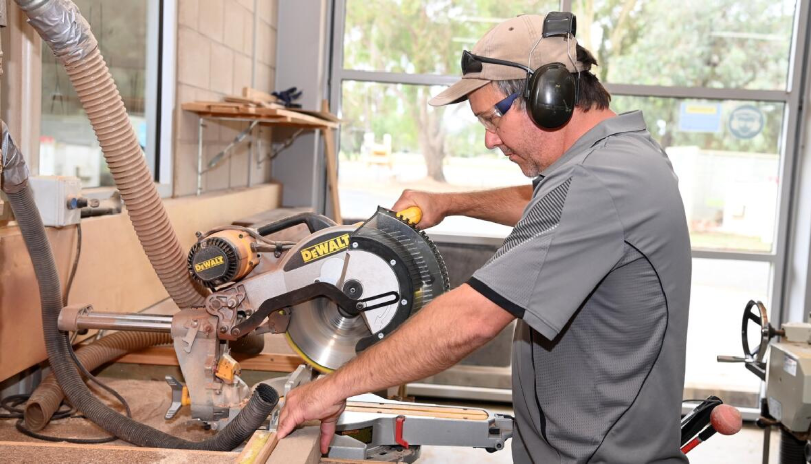 Aaron is using his talking saw to cut a piece of wood. He is wearing a grey polo shirt, a cap, and protective headwear. Photo credit: Shepparton News. Photo by: Rechelle Zammit.