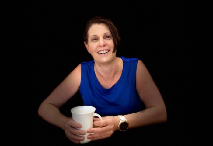 Sarnya is sitting while smiling to camera, wearing a blue top and holding a white mug.