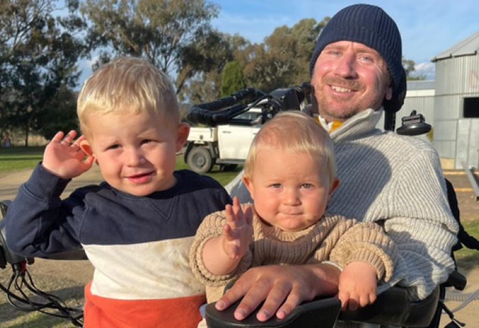 Nathan Stapleton is in a wheelchair outdoors at a farm with his two sons sitting on his lap.