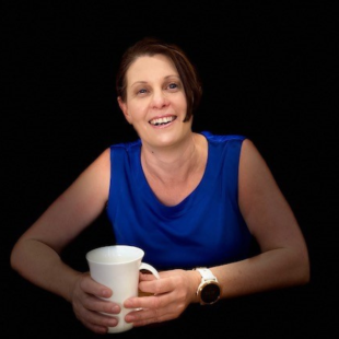 Sarnya is sitting while smiling to camera, wearing a blue top and holding a white mug.