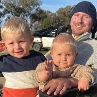 Nathan Stapleton is in a wheelchair outdoors at a farm with his two sons sitting on his lap.