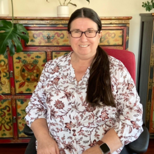 Sharon is smiling facing the camera, sitting in her red and black office chair. She is wearing a floral-patterned top.